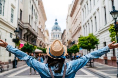 Person with arms raised wearing a hat and denim jacket on a city street with historic buildings.