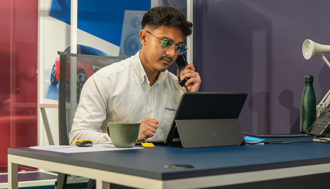 Man on phone working on laptop at office desk with cup and lamp.