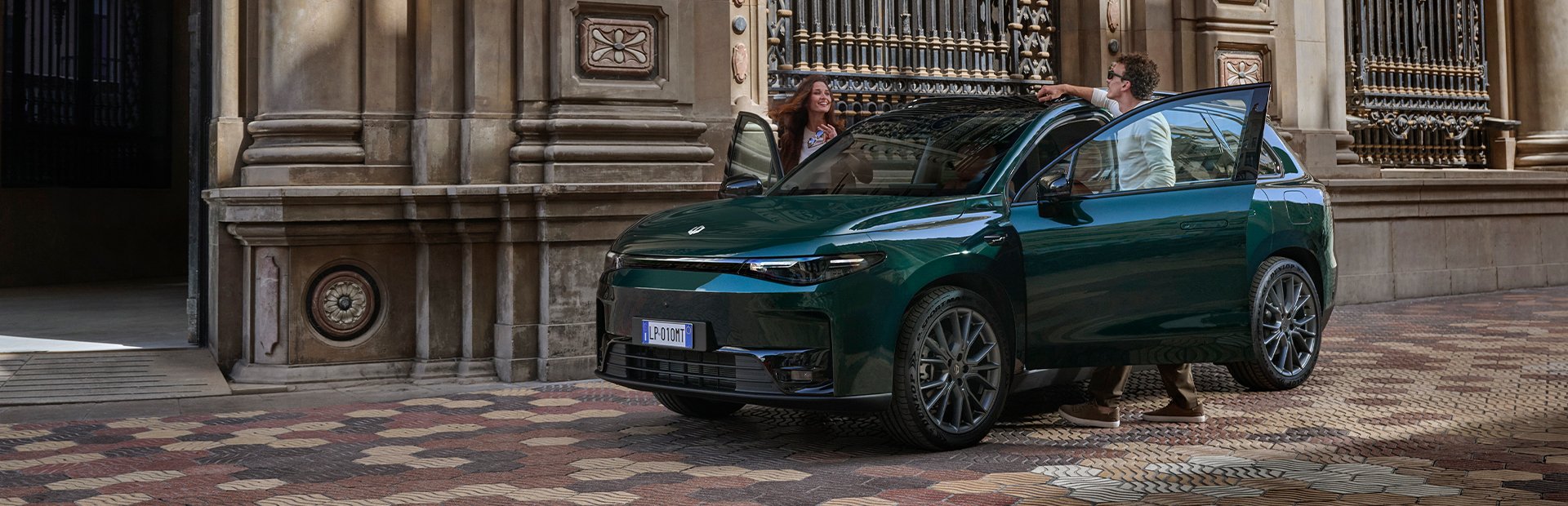 Two people by an open door of a green SUV parked next to a historic building.
